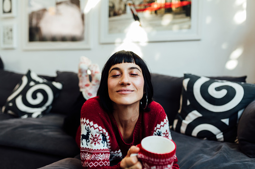 Happy young Caucasian woman celebrating winter holidays at home alone, smiling and enjoying a cup of hot coco