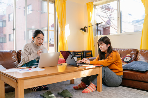 Two young women are sharing an apartment together and doing their own things at home. One woman is working on a laptop and other university student is reading a book while using a laptop.