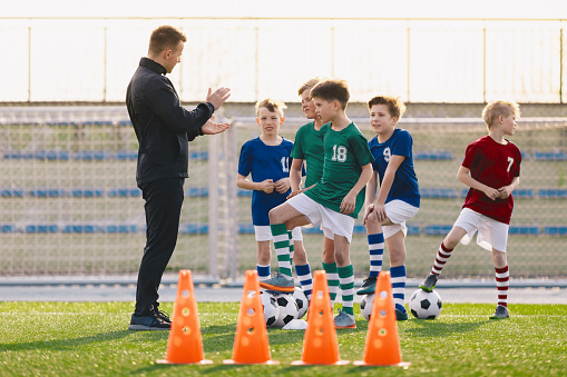 Group of young boys on football training. Kids practicing soccer on grass field. Young man as a soccer coach explaing to players training rules. Children exercising with soccer balls