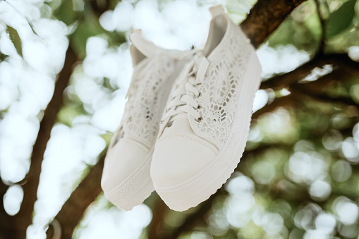 White wedding sneakers hanging on a tree branch. Focused on the front of the sneaker. In the background blurred branches and leaves of trees with a shining arc in the form of defocused bokeh.