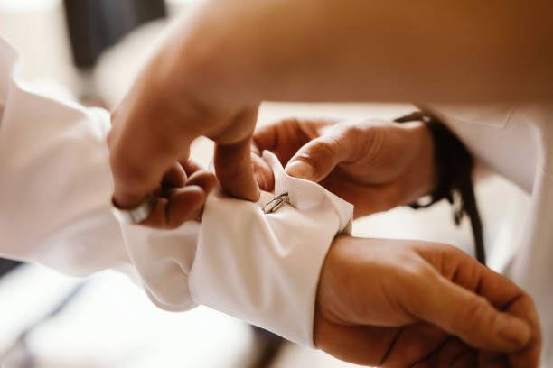 Male hands fastening cufflink at sleeve of white shirt Close-up of male hands (3 hands) fastening cufflink at sleeve of white shirt in helping each other. The scene is in bright light with shallow depth of field. Focused on the cufflink. cufflink stock pictures, royalty-free photos & images