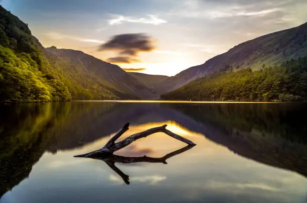 A tranquil scene of a lake between mountains and woodlands with a log in the foreground at sunset