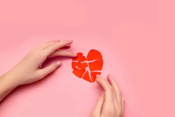 Female hands gluing together broken heart, pieces of torn red valentine on pink background. Concept of disappointment in love, salvation of relationships. Close-up photo