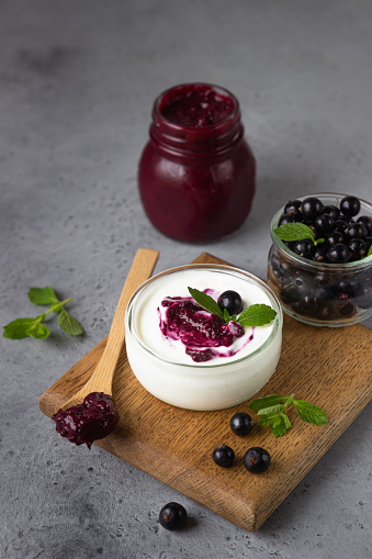 Jar of delicious homemade black currant curd, custard or jam with natural yogurt and fresh berries on grey concrete background. Healthy breakfast.