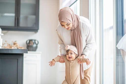 A Muslim mother is encouraging her baby girl to learn how to walk. She is holding her daughter by her arms and attempting to help her stand on her feet.