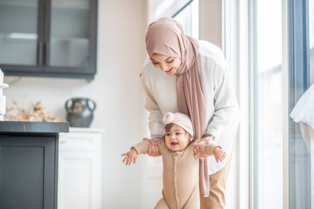 Help me walk, mummy! A Muslim mother is encouraging her baby girl to learn how to walk. She is holding her daughter by her arms and attempting to help her stand on her feet. pics of a cute muslim outfits stock pictures, royalty-free photos & images