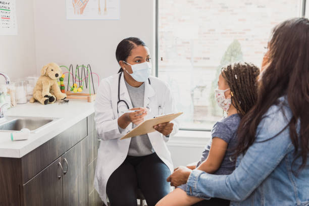 Female pediatrician talks with patient's mother With a protective mask on, a female pediatrician talks to a young patient's mother about the woman's daughter's medical conditions. They are wearing protective masks during the COVID-19 pandemic. pediatrician stock pictures, royalty-free photos & images