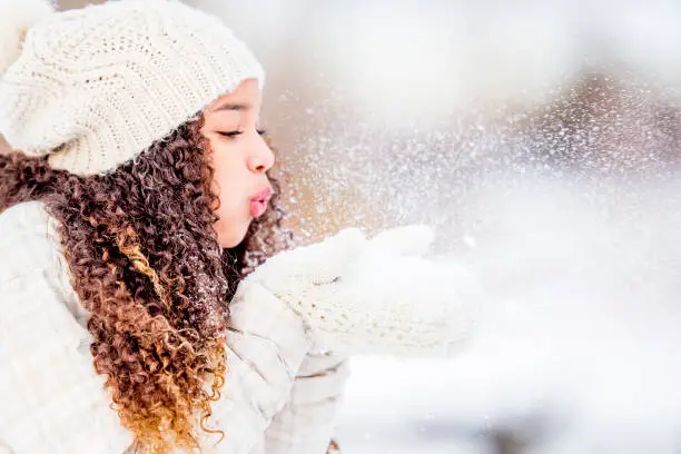 A mixed race female teenager blows snow away from her hands as she enjoys nature in the cold winter time.