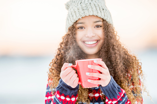 A portrait of a beautiful mixed race female teenager holding a warm mug of coffee outside in the snowy winter weather. She warms a warm sweater and a winter hat.