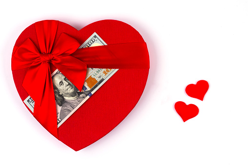 A 100 American dollar bill lies under a ribbon on a gift box in the form of a human heart symbol next to 2 small  symbols of hearts on a white background