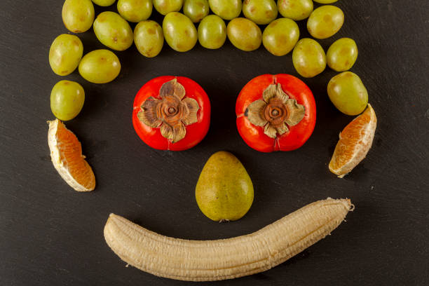 Pear, grapes, persimmon, oranges and peeled banana are placed on a black background to resemble smiling human face.