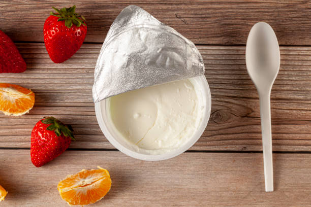 A quick store bought Greek yogurt snack Flat lay image of a store bought plastic cup of plain low fat Greek Yogurt with the aluminum foil seal peeled off. It is on wooden background with a plastic spoon and fresh fruits. A diet idea concept yogurt stock pictures, royalty-free photos & images