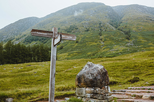 Mount Ben Nevis Landscape Guide board, besides a rock as landmark and climbing path to far away. Background is green grass and trees on Mount slope.