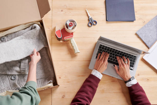 istock Above view of unrecognizable woman packing handmade cardigan into box for dispatch while her colleague typing on laptop 1293702768