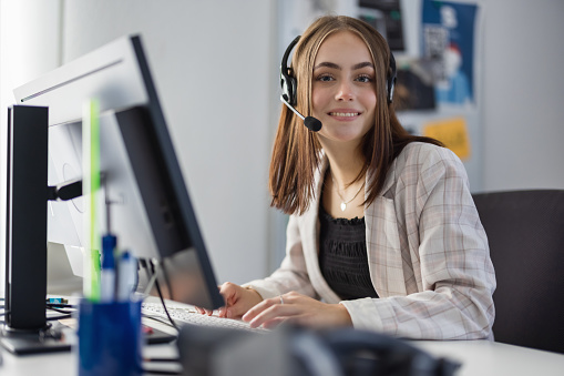 Young businesswoman wearing a headset having a video call on her computer. Sitting inside the Home Office in front of Computer Display looking over towards the camera with a bright smile. Freelance - Female Modern Home Office Teleworking Concept.