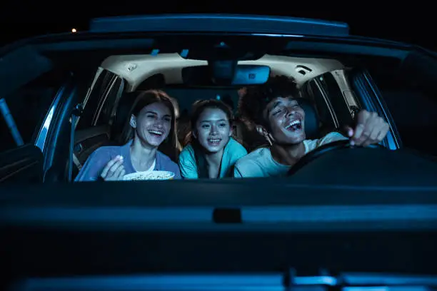 Portrait of three young friends looking emotional, laughing while sitting together in the car and watching a movie in a drive in cinema. Entertainment, leisure activities concept. Horizontal shot