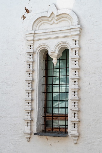 A fragment of a white stone wall of an old building, a narrow window with an iron lattice, stucco decorations, a traditional Russian pattern, arches, decorations, functionality, background, details and elements of architecture, summer, daylight.