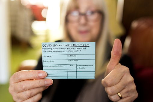 Close up color image depicting a senior woman holding a card with details of her covid-19 vaccination. The woman is happy and positive and is giving a thumbs up gesture. Focus on the card in the foreground while the woman is defocused beyond. Room for copy space.