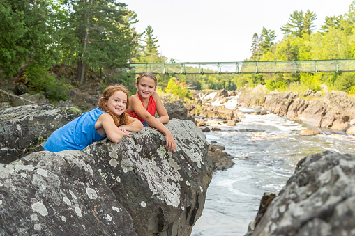 Two sisters on a rocky ledge on the bank of the Saint Louis River at Jay Cooke State Park in Minnesota, USA. The girls are smiling at the camera.
