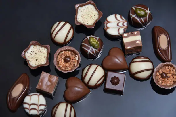 Close up of a selection of luxury chocolates, with a variety of shapes including a heart, florentines, and pistachios. They are in white, milk and dark chocolate and could be for a gift. They are against a grey black background.