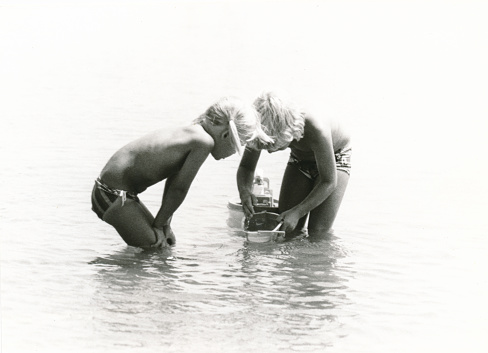 1977 vintage, seventies, retro monochrome image of two young children playing with toy boats in the Mediterranean sea in the French coastal village Bretignolles-sur-Mer.