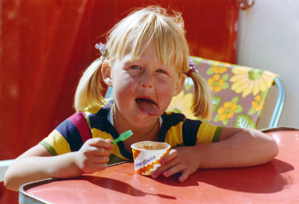 Young girl eating an ice cream with a small spoon Vintage colourful 1977 image of a young girl with striped t-shirt seated on a folding chair at a red formica table eating a ´creme glacée´ (english:´ice cream´) with a small spoon and licking her lips in the French coastal town Bretignolles-sur-Mer. french riviera photos stock pictures, royalty-free photos & images