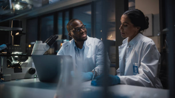 istock Modern Medical Research Laboratory: Caucasian and Black Scientists Work on Laptop, Do Data Analysis, Talk. Advanced Scientific Pharmaceutical Lab for Medicine, Biotechnology Development. Evening Time 1293772872