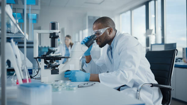 laboratorio de desarrollo médico: retrato de científico masculino negro mirando bajo microscopio, analizando la muestra de petri dish. profesionales que realizan investigación en advanced scientific lab. side view shot - bioquímica fotografías e imágenes de stock