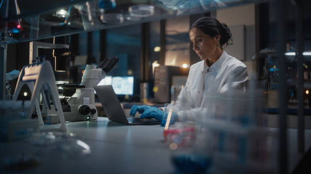 medical development laboratory: female scientist works on laptop, enters data for further analysis. pharmaceutical lab for medicine, biotechnology, bio chemistry, drugs, vaccine research. evening work - laboratory doctor white collar worker research imagens e fotografias de stock