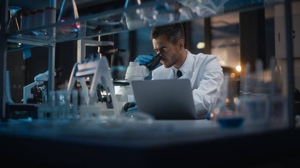 istock Medical Development Laboratory: Portrait of Male Scientist Works on Microscope, Using Laptop, Enters Data for Analysis. Pharmaceutical Lab for Medicine, Biotechnology, Drugs, Research. Evening Work 1293772878