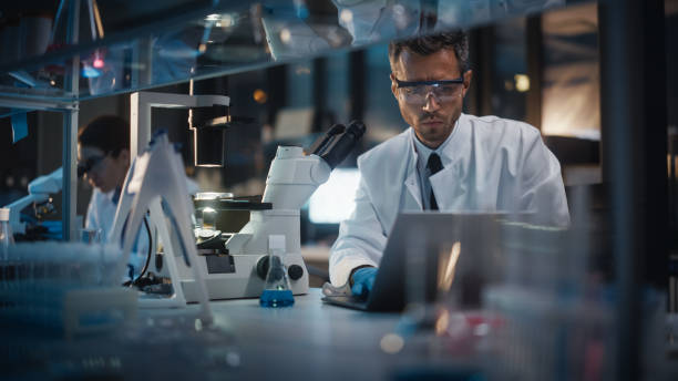 nowoczesne laboratorium badań medycznych: naukowcy płci męskiej i żeńskiej pracują nad laptopem, do analizy danych, użyj mikroskopu. advanced pharmaceutical lab for medicine, biotechnology development. wieczór - medical research zdjęcia i obrazy z banku zdjęć