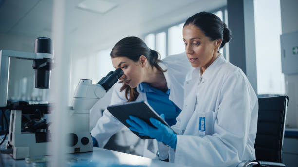 istock Modern Medical Research Laboratory: Two Female Scientists Working Together Using Microscope, Analyzing Samples, Talking. Advanced Scientific Pharmaceutical Lab for Medicine, Biotechnology Development 1293772954