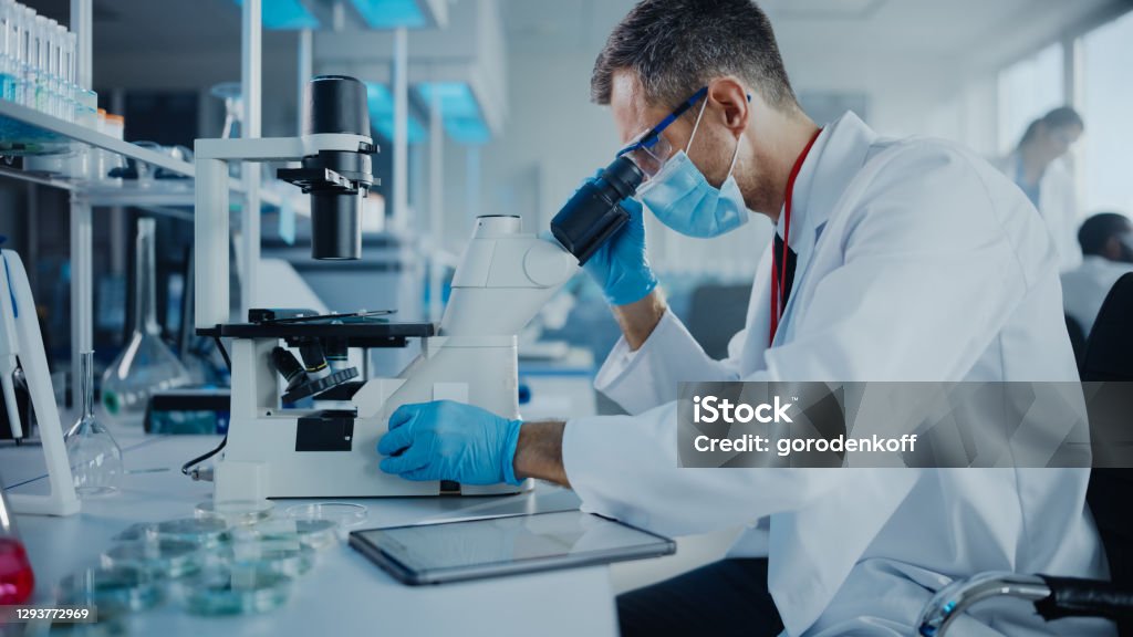 Medical Development Laboratory: Scientist Wearing Face Mask Looking Under Microscope and Using Digital Tablet. Specialists Working on Medicine, Biotechnology Research in Advanced Pharma Lab Laboratory Stock Photo