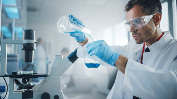 医療開発研究所:男性科学者はガラス用ビーカーに液体化学物質を混ぜて実験を行う。専門医とのバックグラウンド製薬研究室で、バイオテクノロジー - scientist research group of people analyzing ストックフォトと画像