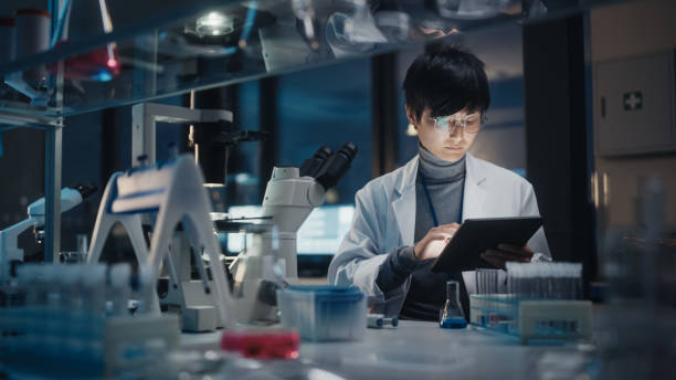 istock Medical Development Laboratory: East Asian Scientist Uses Digital Tablet Computer, Uses Microscope, Conducting Experiment. Pharmaceutical Lab for Research Medicine, Biotechnology. Evening Work 1293773012