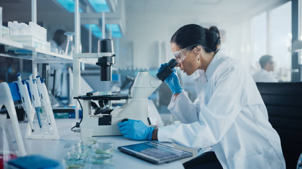 istock Medical Development Laboratory: Caucasian Female Scientist Looking Under Microscope, Analyzes Petri Dish Sample. Specialists Working on Medicine, Biotechnology Research in Advanced Pharma Lab 1293773560