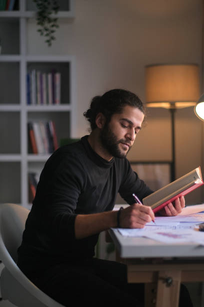 jovem sentado à mesa trabalhando e lendo um livro em casa. - report reading business book - fotografias e filmes do acervo