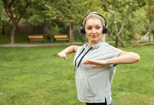 Young fit blonde woman warms up before training and listens to music on headphones in the park. Aerobica
