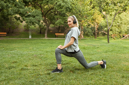 Young fit blonde woman warms up before training and listens to music on headphones in the park. Aerobica