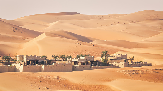 Panorama of Desert Oasis Village with planted palm trees inside the Empty Quarter Desert of Abu Dhabi surrounded with large Sand Dunes. Rub' al Khali Oasis Village under blue cloudless sky. Rub' al Khali Desert and Sand Dunes close to the border of Saudi Arabia and the United Arab Emirates. Abu Dhabi, United Arab Emirates, Middle East, Persian Gulf Countries, Western Asia.