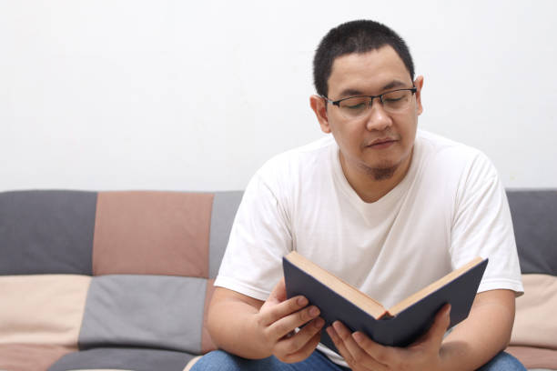 istock Happy Asian man reading a book while relaxing on sofa, enjoy weekend 1293806572
