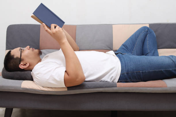 istock Happy Asian man reading a book while relaxing on sofa, enjoy weekend 1293806575