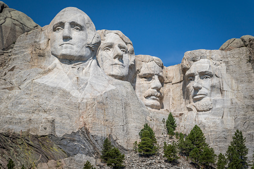 Front view of Mount Rushmore. The carved faces of the four historical figures, George Washington, Thomas Jefferson, Theodore Roosevelt and Abraham Lincoln are framed by a brilliant stormy sky and tree branches. Bright green coniferous trees in the foreground provide contrast to the granite stone.