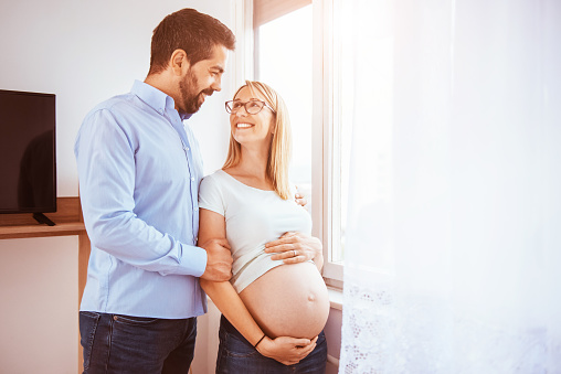 Happy young couple expecting baby, standing together near the window at home