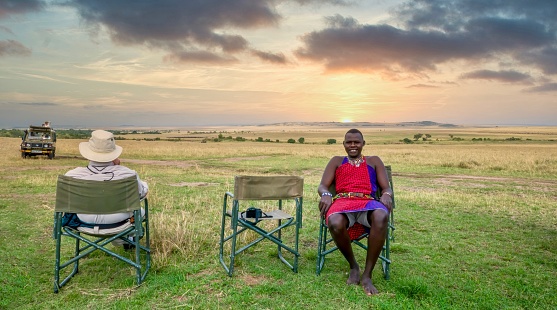 Maasai Mara National Reserve, Kenya - September 29, 2013. A Kenyan safari guide and an American male guest relax on a game drive rest break, as the sun sets over the vast African landscape.