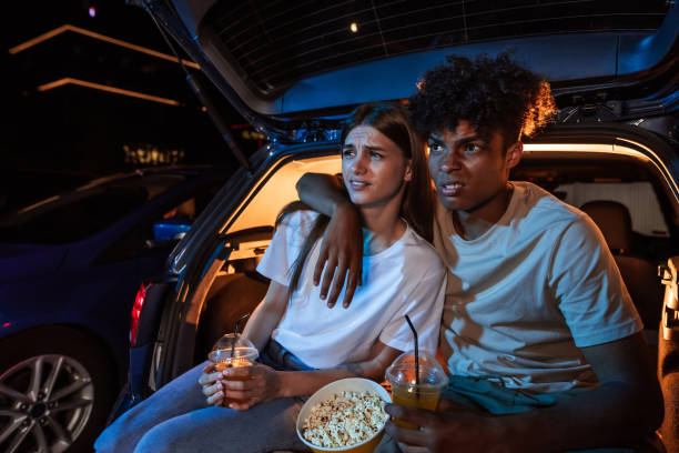 아름다운 젊은 부부는 영화를보고, 야외 영화관에서 큰 화면 앞에서 자동차 트렁크에 함께 앉아있는 동안 팝콘을 가지고 - drive in movie car inside of billboard 뉴스 사진 이미지