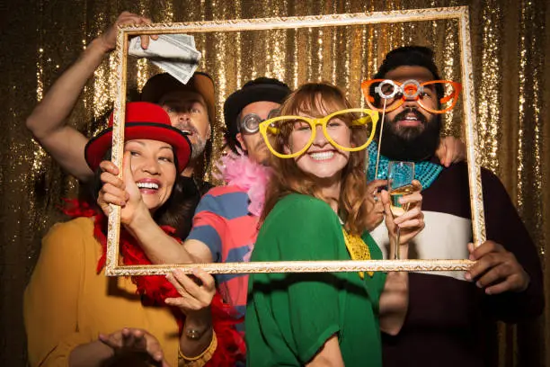 Portrait of cheerful male and female friends wearing party props and taking funny photo with picture frame in photo booth.
