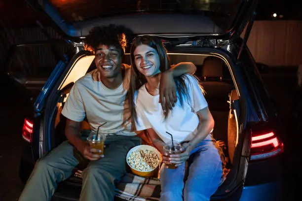 Diverse young couple having romantic date. Cheerful guy and his girlfriend watching a movie, sitting together in car trunk in front of screen in open air cinema. Entertainment ideas. Horizontal shot