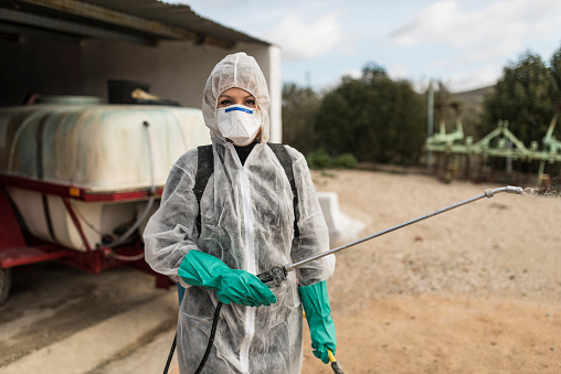 Woman with the fumigation suit to apply the pesticides to the olives. Woman dressed in phytosanitary suit and mask poses with tank for spraying or disinfectant in the background