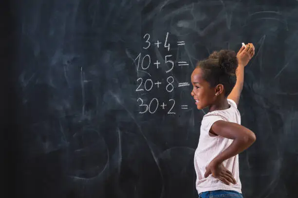 Photo of Mixed race elementary school girl solving math problems on chalkboard